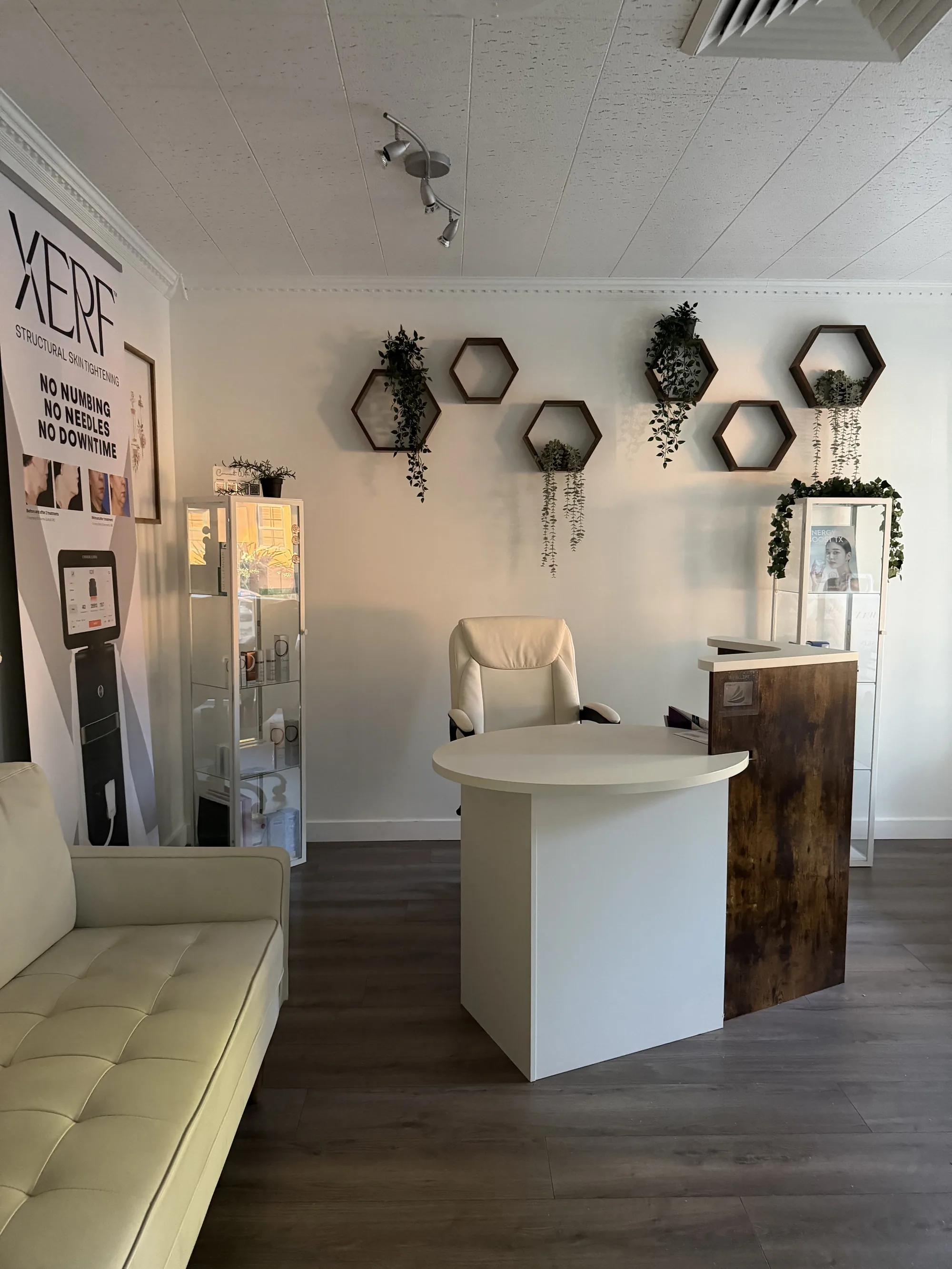 A bright clinic room with a cream sofa, a round white desk, glass display cabinets, hexagon wall shelves with plants and a XERF banner