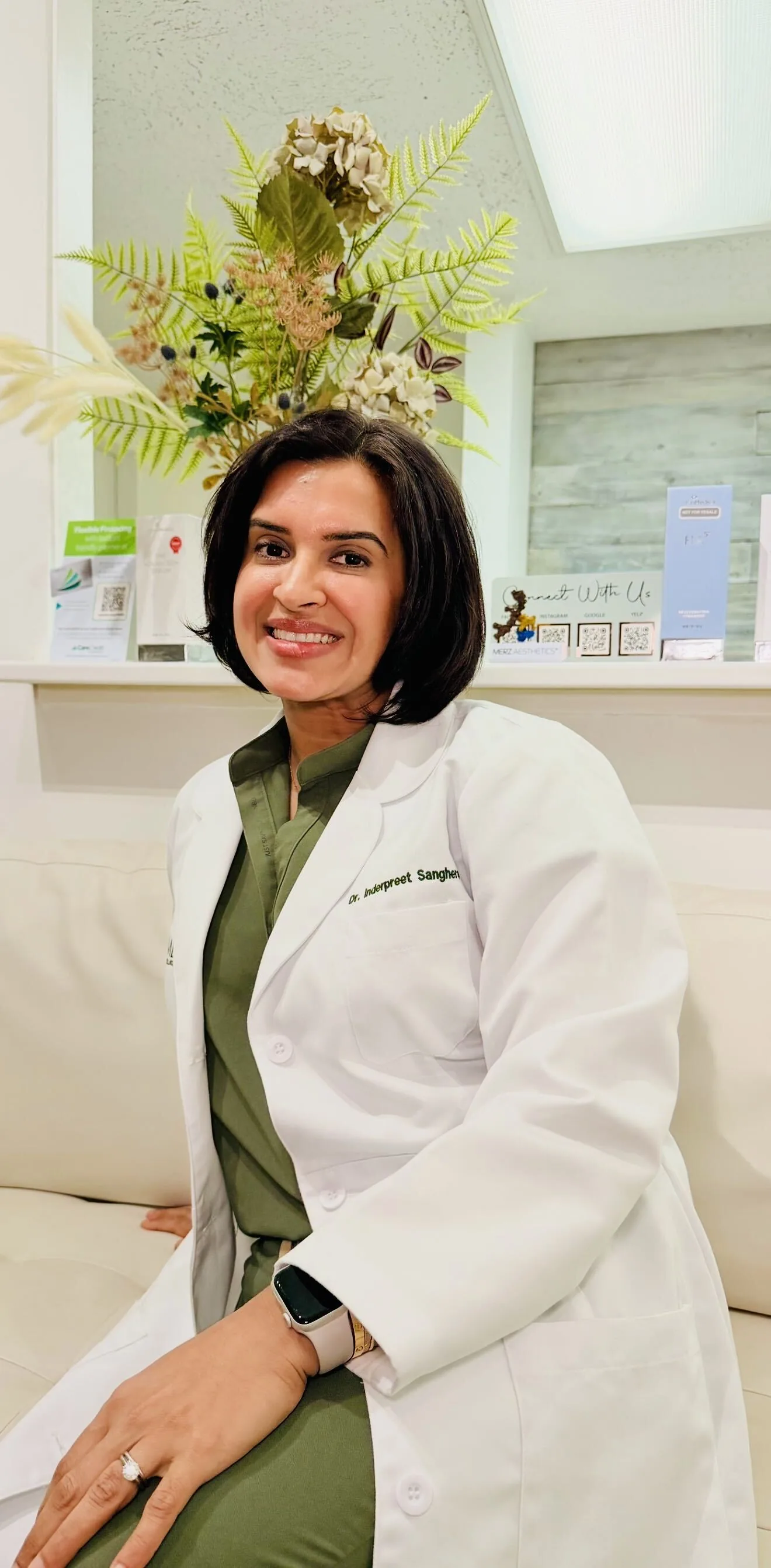 Dr. Inderpreet Sanghera smiling, seated in a white coat and olive scrubs in the clinic