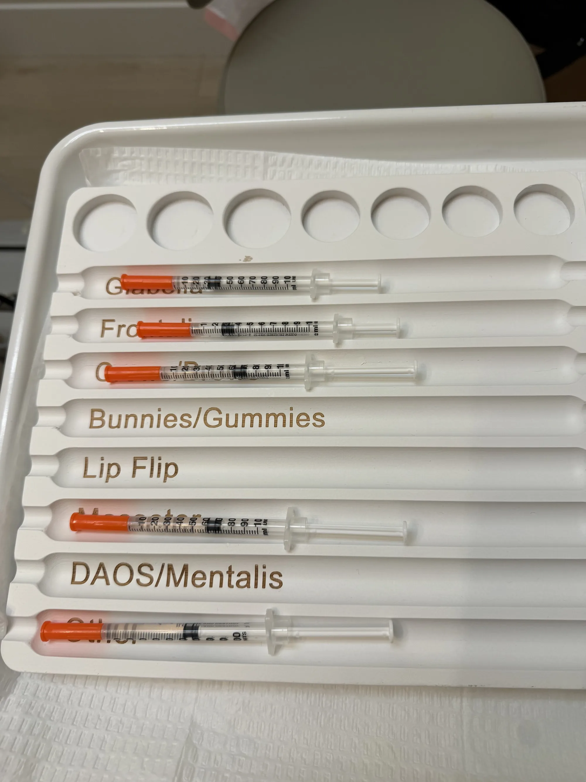 A white tray of small syringes, each slot labelled by treatment area, including bunnies and gummies, lip flip and DAOs and mentalis