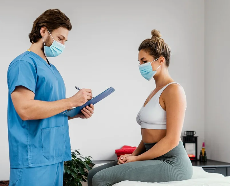 A clinician in blue scrubs and a face mask taking notes on a clipboard while talking with a woman seated on an exam table